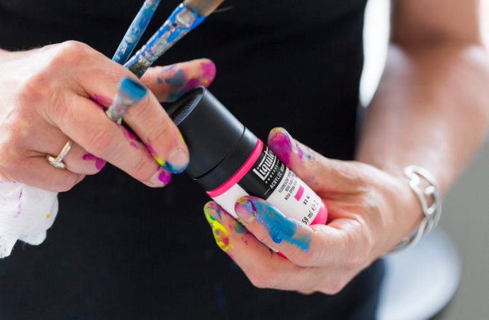 Gros plan sur les mains d’un artiste tenant un flacon de gouache acrylique Liquitex couleur Rose Opéra Fluorescent, avec les doigts et les pinceaux couverts de peinture, illustrant une activité artistique vibrante.