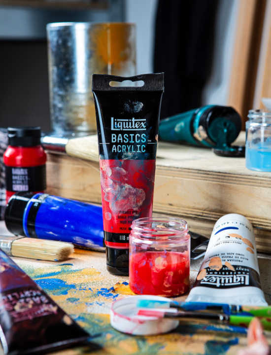 Tube de peinture acrylique Liquitex BASICS couleur rouge posé sur une table d’atelier, entouré de pinceaux, de pots de peinture ouverts, et d’autres tubes de différentes couleurs, dans un environnement artistique vivant et créatif.